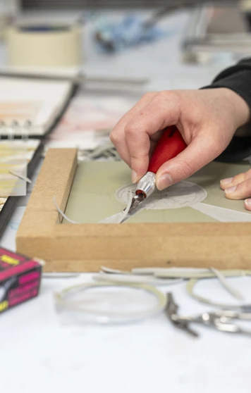 Close-up of hands carving a design into a printmaking block on a desk, surrounded by sketches and art supplies Close-up of hands carving a design into a printmaking block on a desk, surrounded by sketches and art supplies
